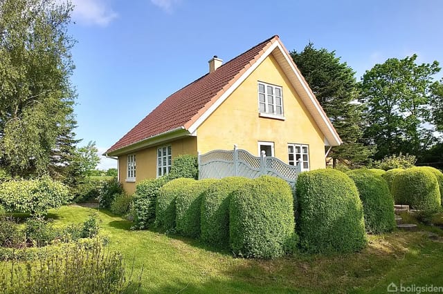 The house stands quietly, surrounded by a well-kept garden with bushes and trees. It has a yellow facade, red tile roof, and several windows, giving it a cozy appearance in the green surroundings.