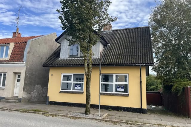 Yellow house with black roof, 'For Sale' signs in the windows, located along a street with a tree in front. Neighboring houses have different colors and roof designs.