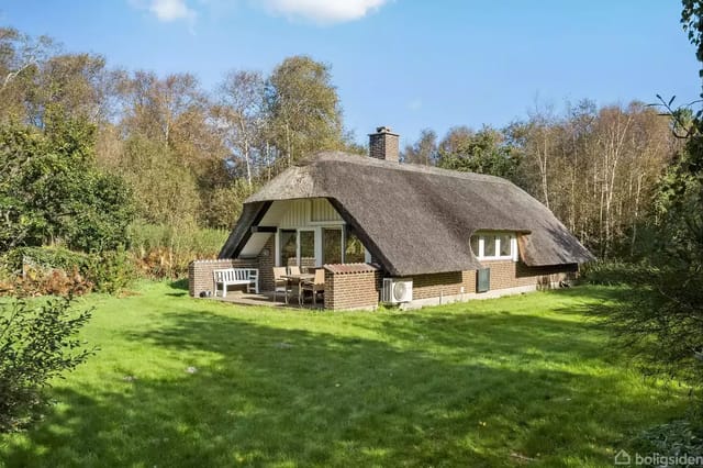 Thatched house in a green garden surrounded by trees. The terrace is furnished with a garden table and chairs.