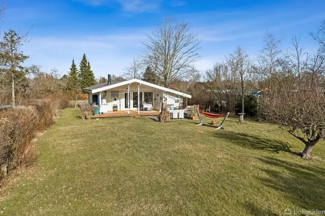 A small holiday home stands in a green garden with a hammock to the right. Trees and bushes surround the area under a blue sky.