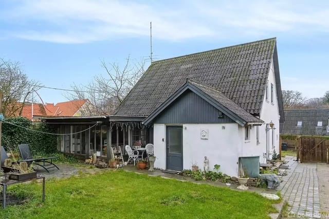 A white house with a gray roof stands in a garden with a green lawn. In the foreground, there are garden furniture and potted plants. The house has a veranda with a glass roof.