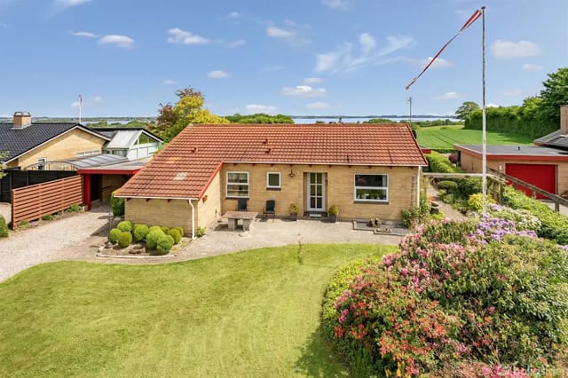 A yellow brick house with a red tiled roof, surrounded by a well-kept garden with grass, bushes, and flowers. To the left is a gravel path and to the right a flagpole.