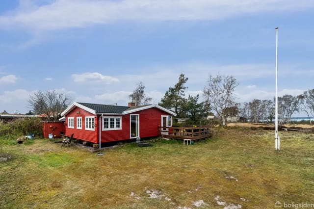 Red wooden house on an open plot surrounded by grass and scattered trees, with a flagpole and a clear sky.