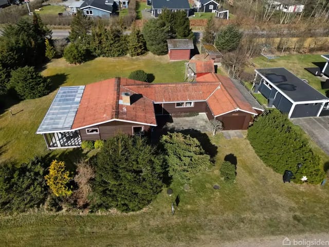 A detached house with a red roof and garage surrounded by a garden with bushes and trees. Other houses are visible along the street in the background.