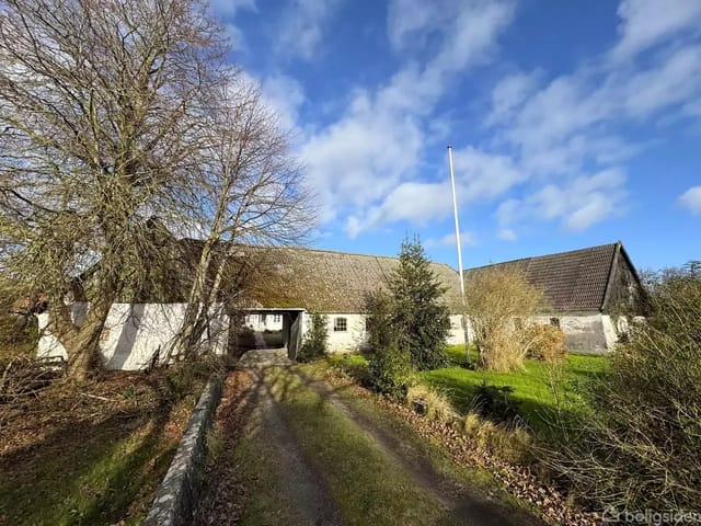 A thatched farmhouse surrounded by trees and green bushes under a clear blue sky. A gravel path leads to the main entrance.