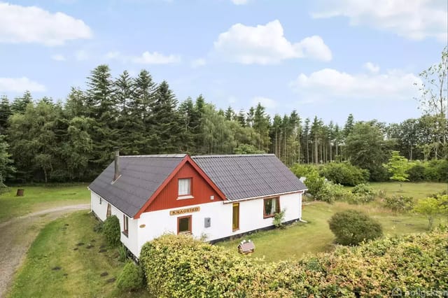 A white house with a red roof stands quietly in a lush, wooded garden. The sign 'Enkekilden' is visible on the facade.