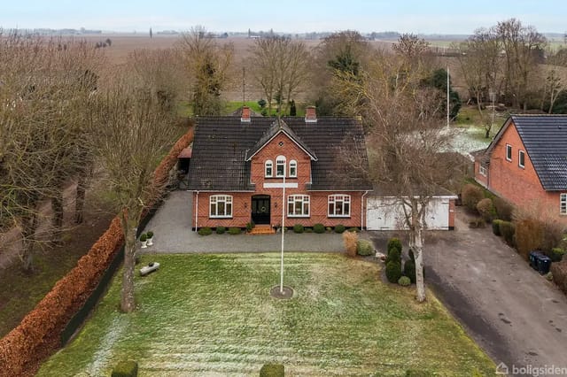 Red brick house with dark roof in a well-kept garden, surrounded by bare trees and neighboring houses. The grass shows traces of frost, and there is a paved driveway.