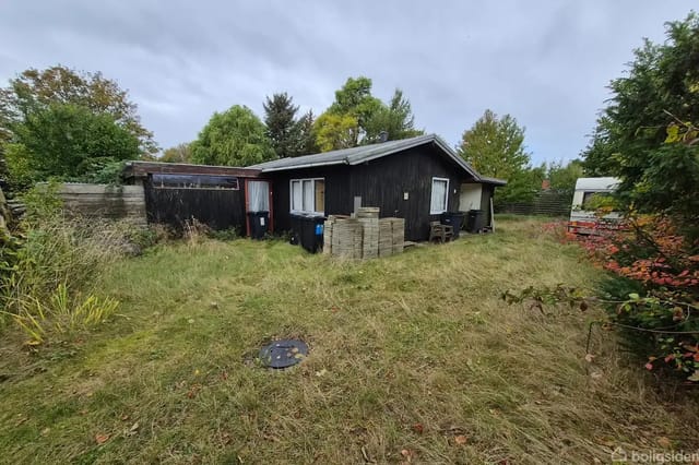 A black wooden building stands quietly surrounded by a lawn with tall grass and bushes, under a gray sky. In the background, there are more trees and some outdoor items.
