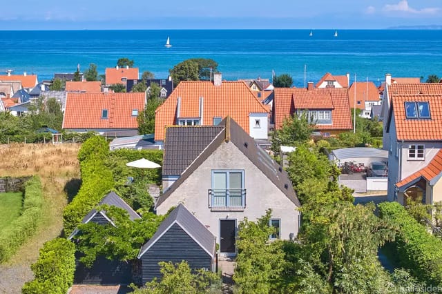 Gray house with sloped roof, surrounded by green foliage in a coastal town with red roofs. The sea and boats are visible in the background, creating a summery atmosphere.
