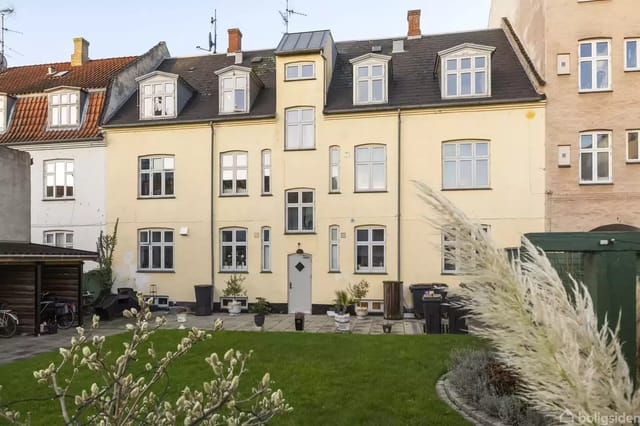 A yellow townhouse with a sloping roof and several windows stands in the middle of a green, well-maintained garden area. Surrounding buildings complement the urban atmosphere.