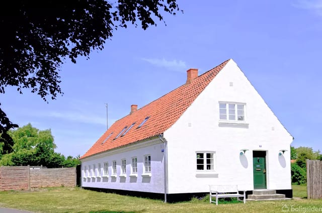 A white, elongated house with a red tile roof on a lawn under a blue sky. The house has several small windows and a green front door. A bench stands in front of the entrance.