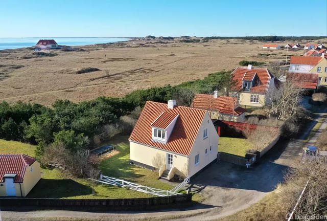 House with red tile roof on a lawn surrounded by a white fence. It is located in an open landscape near the beach and dunes, with several houses nearby.