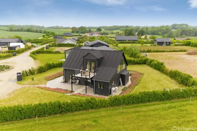 A modern black wooden house with a raised terrace stands in a well-kept garden, surrounded by green fields and a few similar houses in the background, creating a peaceful landscape feeling.