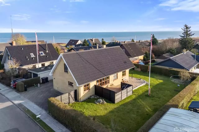 A two-story house with a dark roof on a well-maintained lawn with a hedge around. In the background, a row of villas and sea view under a clear blue sky.