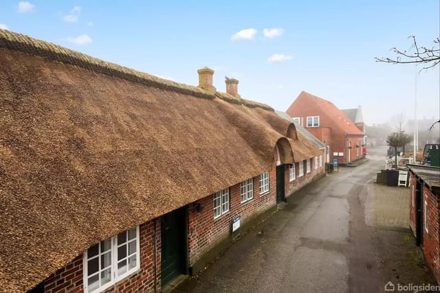 Thatched half-timbered house with red bricks along a quiet street in a town. The house has white windows, and a larger red building is seen further down the road.