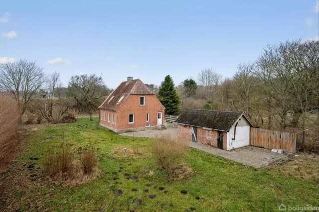 A red brick house on a green plot with thin trees and bushes. Next to the house is a smaller building with a white door and a black tile roof.