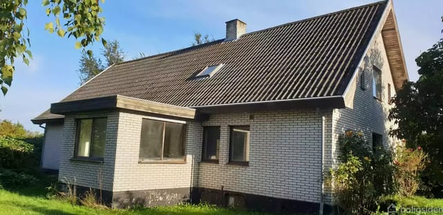 A single-family house with gray brick walls and a black sloped roof has windows facing sunlight. It stands in a garden with green trees and bushes around.