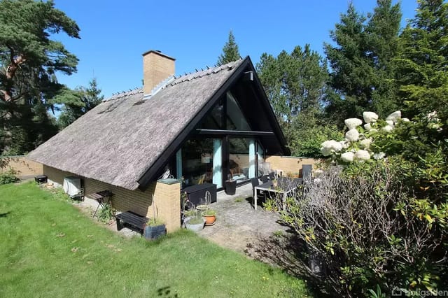 A thatched cottage with large glass sections stands in a lush garden surrounded by trees and bushes. A terrace with potted plants and furniture is seen in front of the house.