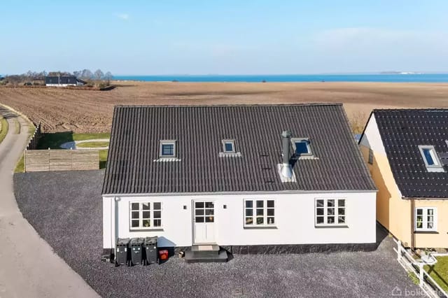 A white house with a black roof stands quietly on a gravel road, surrounded by fields, with a blue horizon, and close to the sea in the background.