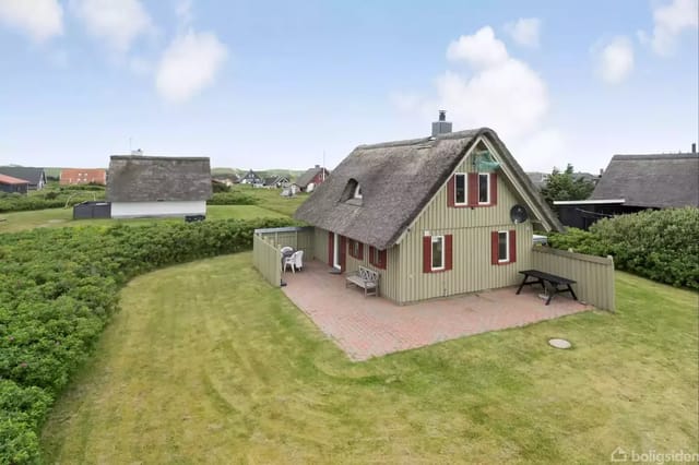 A small thatched house stands on a well-kept lawn with a terrace and garden furniture, surrounded by other houses and green bushes under a blue sky with clouds.