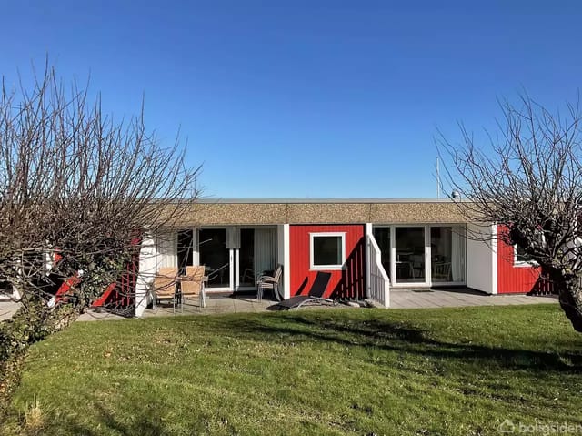 A red and white house with a flat roof stands in sunny weather. In front of the house is a lawn and pruned trees, surrounded by a clear blue sky.