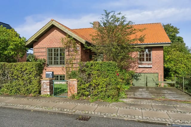 A red brick house with a red tiled roof, surrounded by green bushes and trees. Driveway to the right, sidewalk in front, under a blue sky.