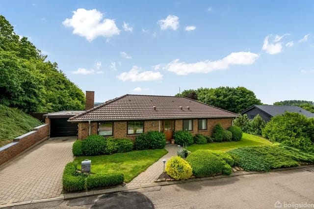 A brick house with a red tile roof stands in the middle of a green garden surrounded by bushes. The house has a paved driveway and a carport. A blue sky with clouds above.