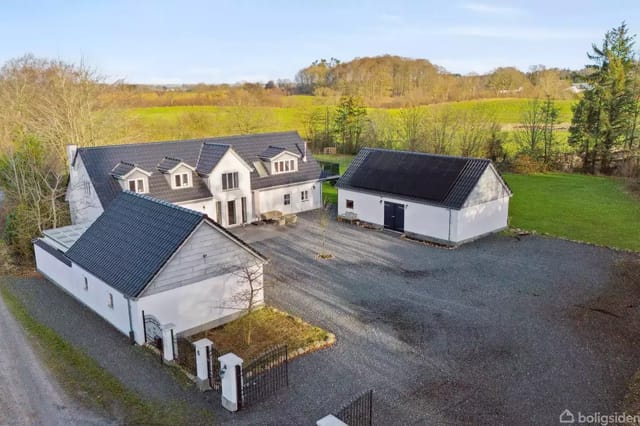 A white building with a black roof stands on a gravel courtyard surrounded by green fields and trees under a blue sky.