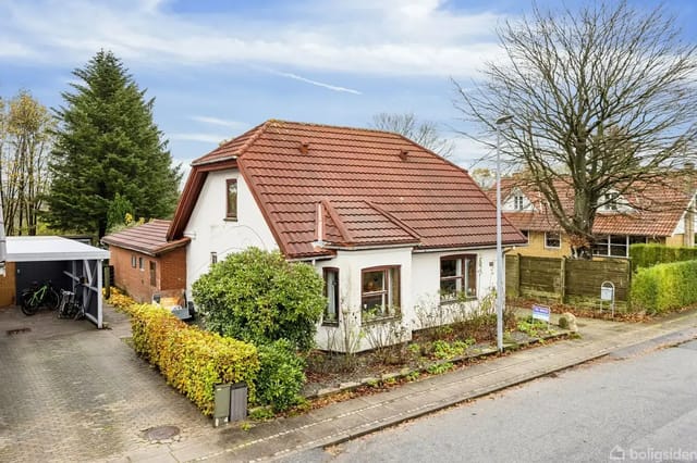 A white house with a red tile roof on a peaceful residential street, surrounded by hedges and trees. A carport to the left houses bicycles. Sign by the road.
