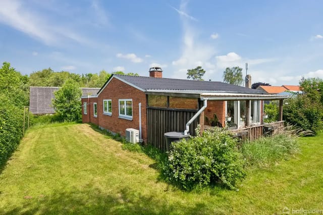 Red brick house with covered terrace in a lush garden under a blue sky with scattered clouds.