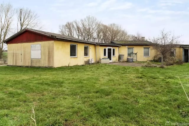 A yellow brick building with a flat roof stands in a grassy area. An abandoned atmosphere with a few trees in the background.