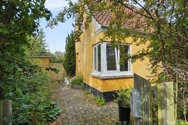 Yellow house with sloping roof in a lush garden, stone path leading to a wooden gate