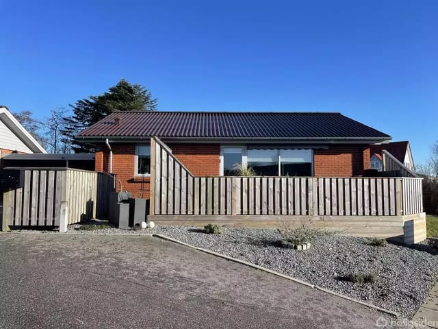 A red brick house with a black roof behind a low fence. In front, there is a gravel driveway with some plants. Clear blue sky in the background.