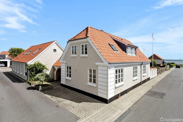 A white house with a red roof stands quietly on a cobbled street beside another similar house. A blue sky and sea view are seen in the background.