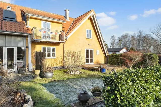 A yellow brick house stands in a garden with a lawn and bushes. A terrace with potted plants is seen to the left. The house has a red tiled roof and a balcony on the first floor.