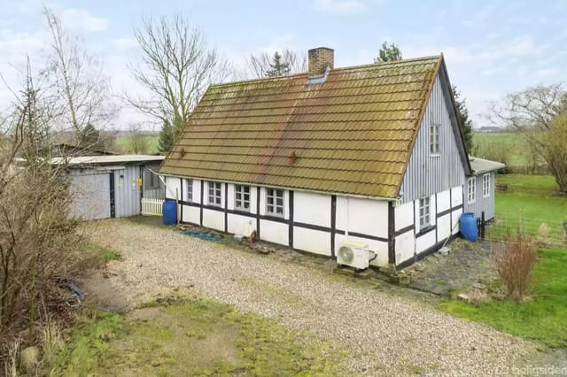 A timber-framed house with a brown tile roof stands on a gravel road. Next to it is a wooden building and grassy area with scattered trees. The atmosphere is rural and open.