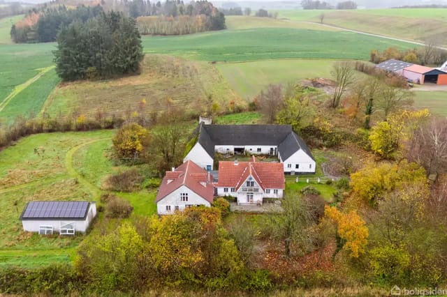 White country house with red roof surrounded by autumn-colored trees, located in a rural area with fields and a nearby barn.