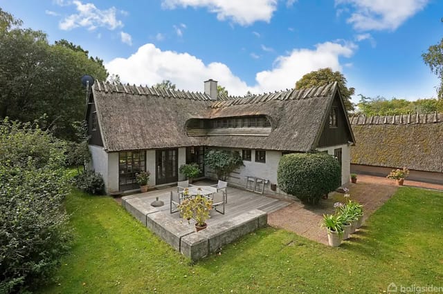 Thatched half-timbered house in a lush garden with terrace, surrounded by bushes and trees under a blue sky.