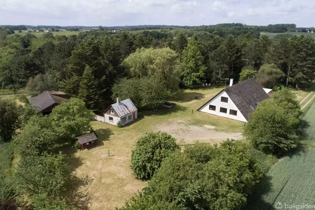 A triangular house with white walls stands quietly in a wooded rural setting, surrounded by green trees and fields under a clear sky.