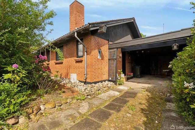 A red brick house with a chimney, set in a garden with flowers and bushes. A paved path leads to a carport beside the house.