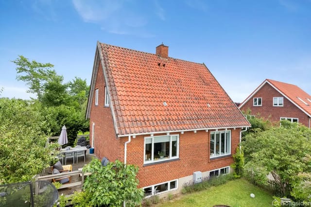 A red brick house with a red tile roof stands in a green garden with an outdoor terrace area and garden furniture. Another similar building is seen in the background under a clear blue sky.