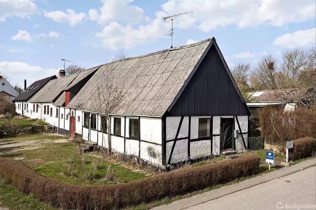 Old half-timbered house in a landscape with gravel roads and small bushes. A real estate agent sign is in the foreground. The house has a large, sloping roof and visible beams on the facade.