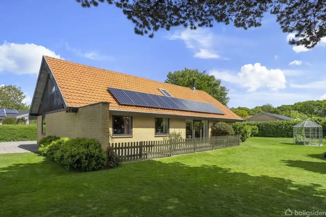 House with red tile roof and solar panels on a lawn surrounded by bushes and trees; a small greenhouse is seen in the background under a clear blue sky.