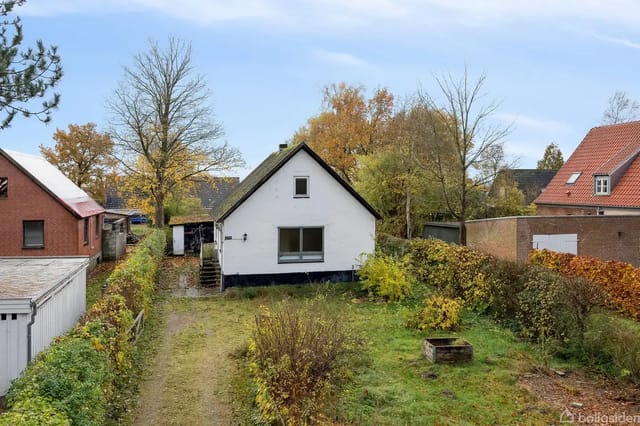 The house is white with a black base and sloped roof, set in a garden with trees and bushes.