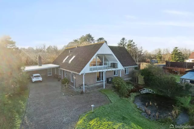 A two-story brick house with a sloped roof stands in the foreground. A white car is parked in a gravel driveway. The house has a balcony, and the surroundings include trees and a small garden with a pond.