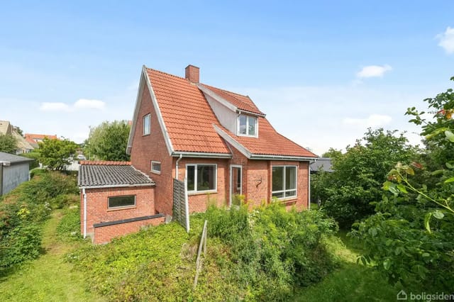 A red brick house with a red tile roof stands quietly in a lush garden with green bushes and grass around. Clear weather with blue sky.