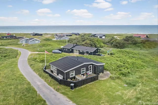 A black summer house with a terrace on a grassy plot near the road. The surroundings include several houses and a sea view in the background under a blue sky with clouds.