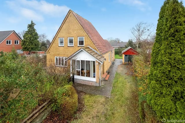 A yellow brick house with a red tile roof stands in a garden with grass and bushes. A smaller red building is seen in the background. The sky is blue with a few clouds.