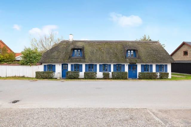 Thatched house with blue shutters on a quiet residential street, surrounded by bushes and trees under a clear blue sky.