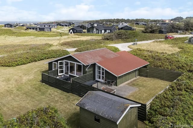 A wooden holiday home with a reddish-brown roof and terrace situated in a green, hilly landscape. Surrounded by other houses and roads in the background under a blue sky.